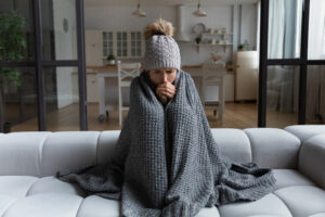 Woman wearing a beanie bundled up on the couch under a blanket because heater is blowing cold air.
